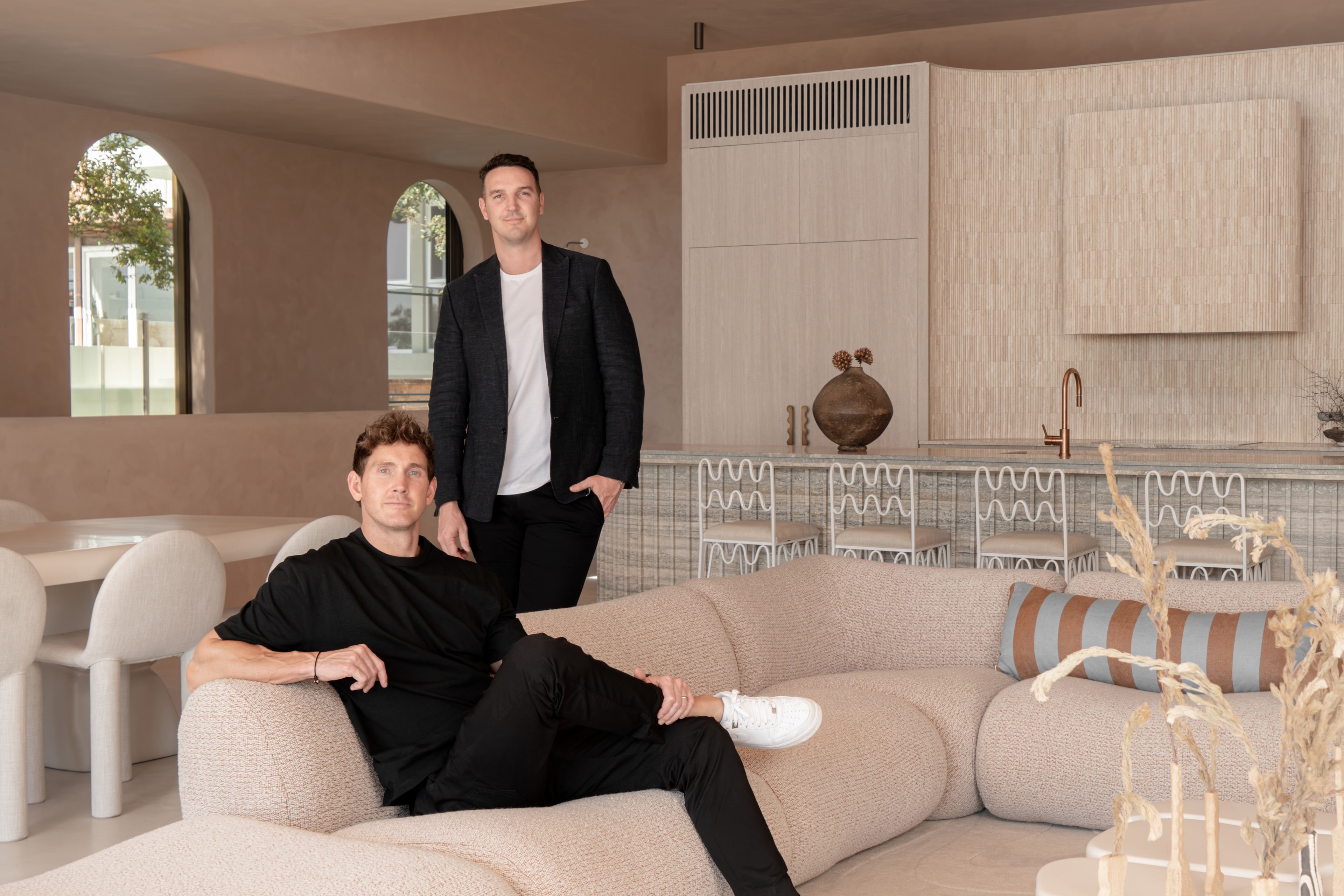 Two founders in a warm-plaster living space with curved sofa and travertine kitchen. Graya